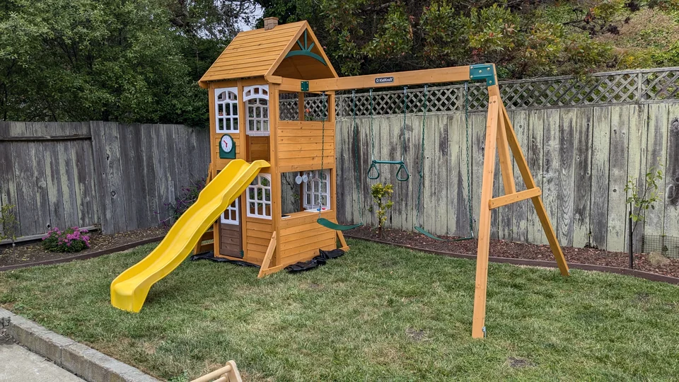 KidKraft wooden playset with playhouse windows, a yellow slide, two swings, and a trapeze, assembled by Artem in June 2024